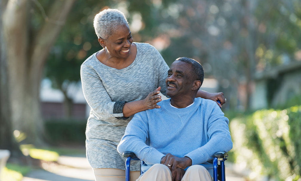 Elderly man sitting in a wheel chair being pushed by a woman.