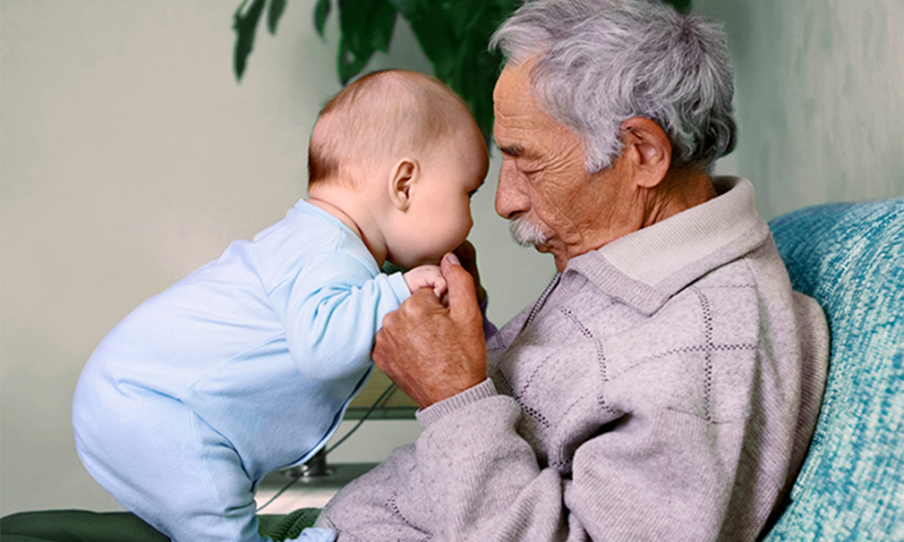 Elderly man wearing a grey sweater holding a baby in a blue onesie.