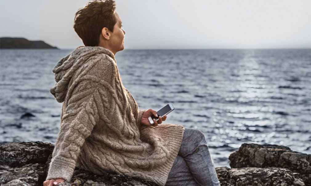 Individual looking out into the ocean holding a cell phone and wearing a tan sweater.