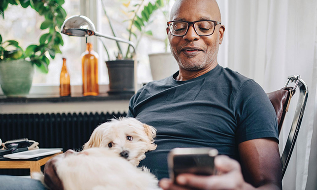 Man sitting down on a couch wearing a grey t-shirt and glasses looking at his phone with a small white dog on his lap.