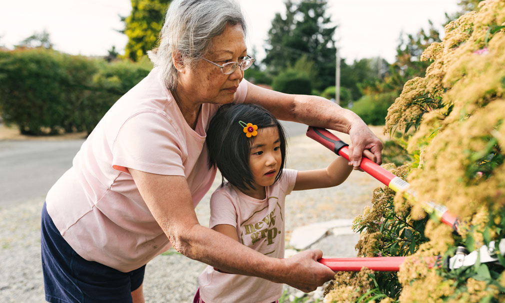 Elderly woman showing a young girl how to cut trees outside.