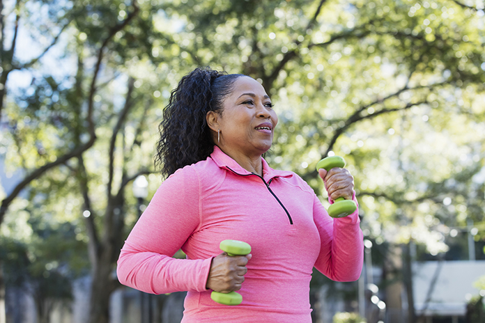 Female wearing a pink top exercising outside with two green weights.