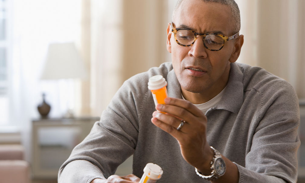 Man wearing a grey sweater and eyeglasses holding a prescription bottle and reading the label on it.
