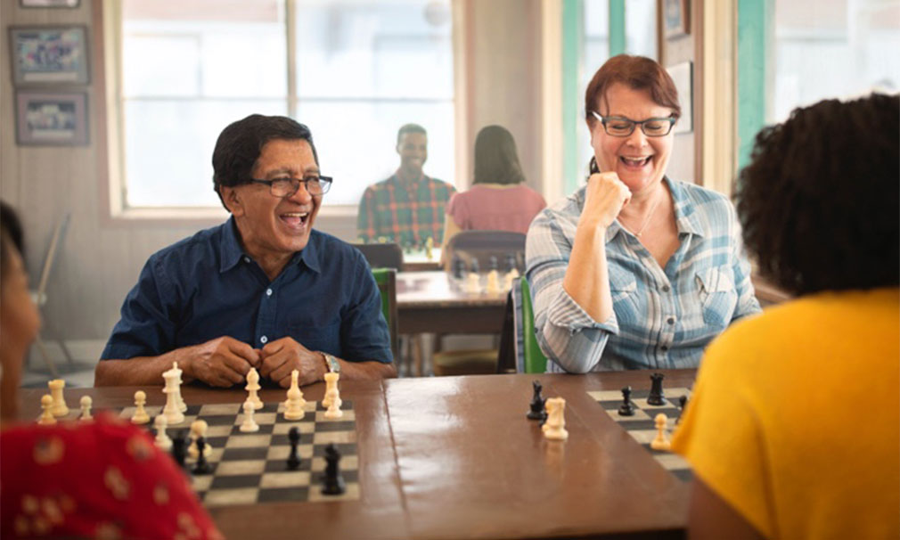 Group of people laughing while sitting down at a wooden table playing chess.
