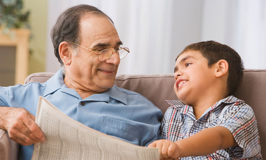 Elderly man wearing glasses and a blue shirt sitting down on a couch speaking to a young boy.
