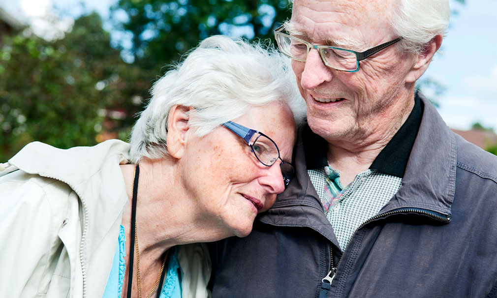 Elderly woman wearing glasses resting her head on an elderly man's shoulder.