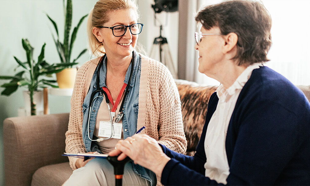 Female healthcare provider wearing eyeglasses and a stethoscope sitting on a couch with a patient.