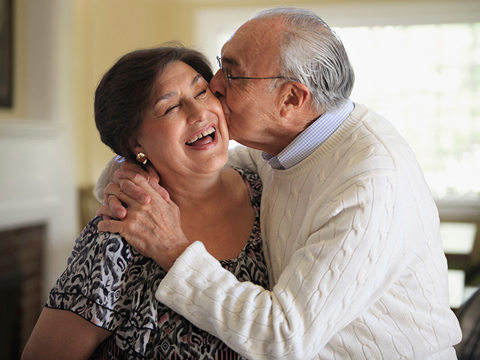 Elderly man kissing a woman on the cheek as she smiles.